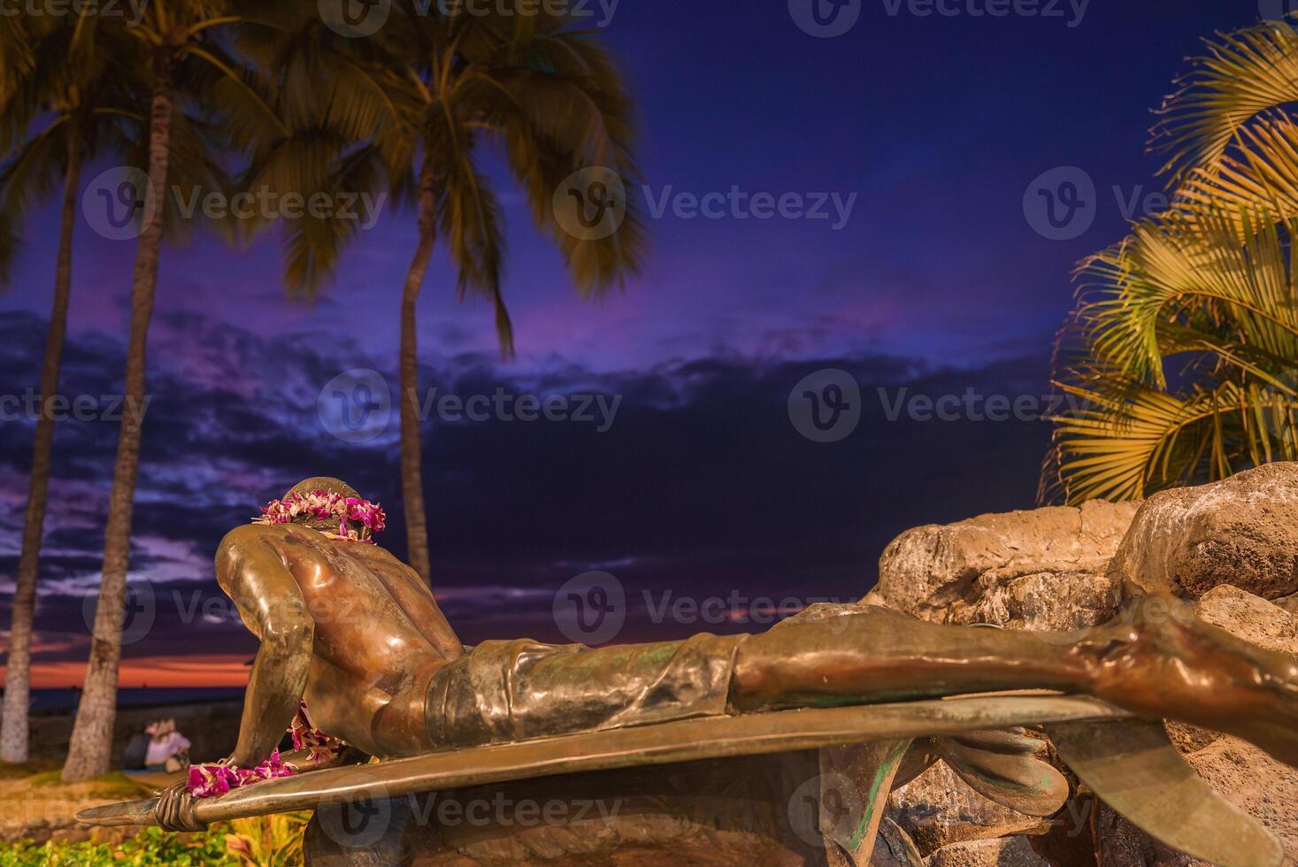 A bronze surfer statue adorned with flower leis lies on a surfboard, framed by palm trees and tropical foliage, set against a vibrant sunset sky. photo