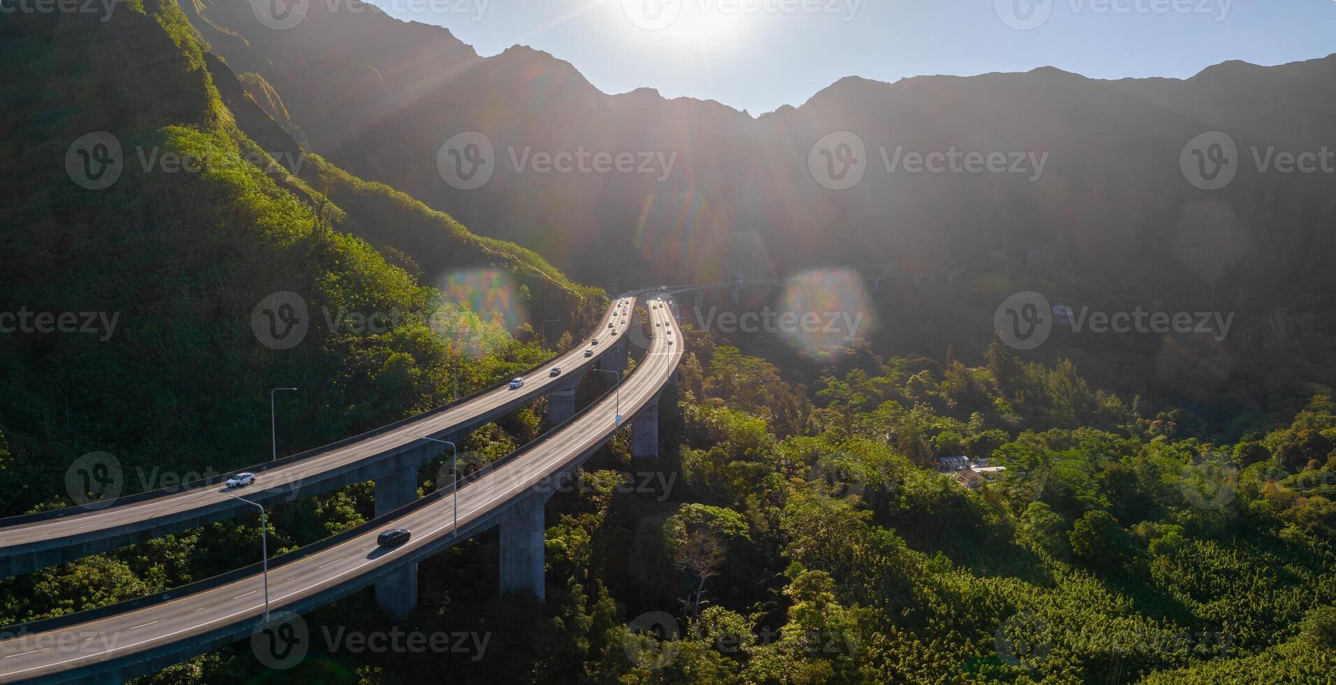 An elevated highway curves through a lush valley on Oahu, Hawaii, surrounded by dense vegetation and steep cliffs under sunlight with lens flare. photo