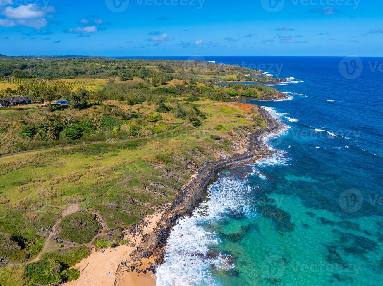 Aerial image of Kauai's coastline showing rugged cliffs, lush greenery, turquoise ocean waters, sandy beaches, and a clear sky with scattered clouds. photo