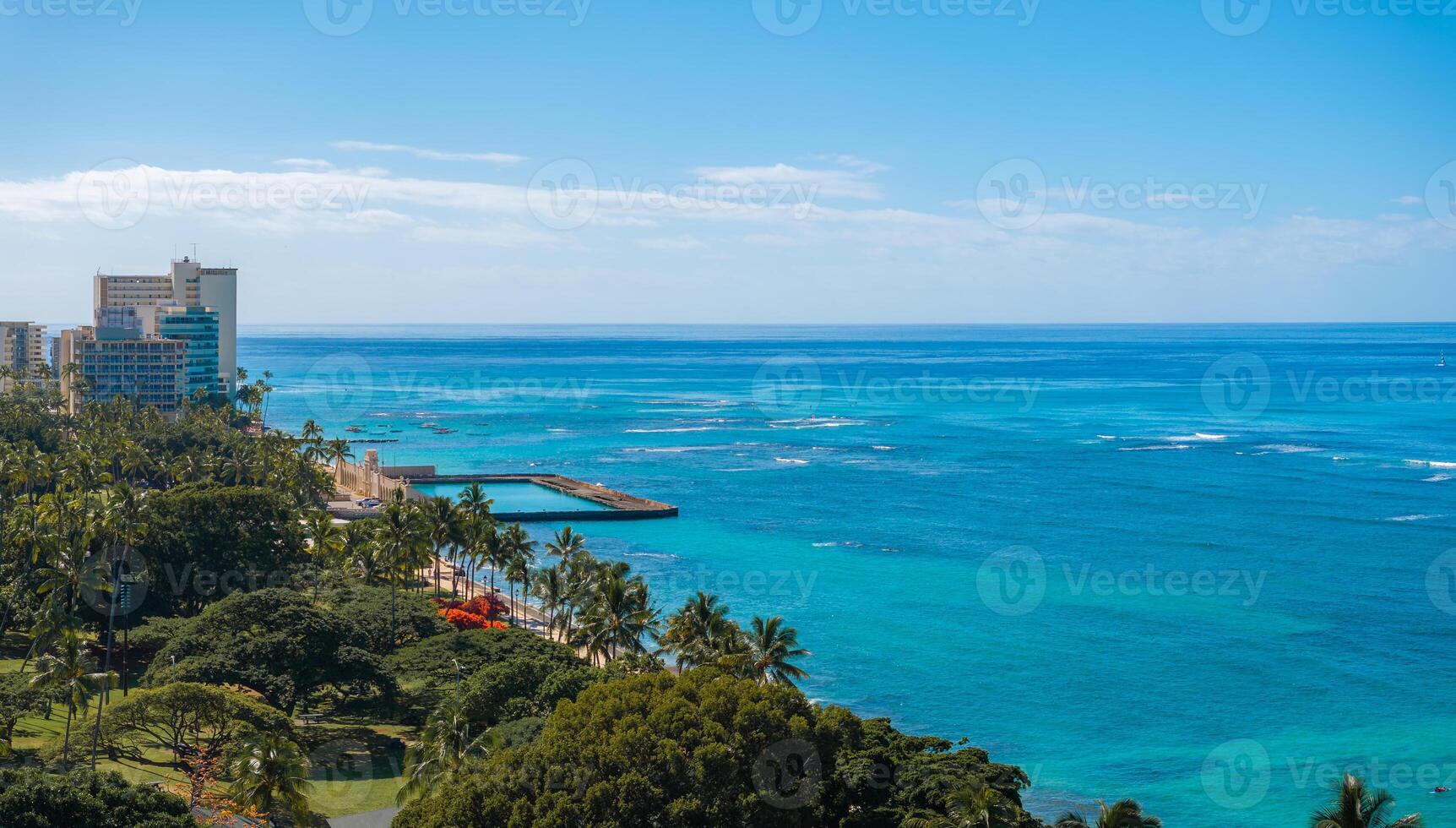Aerial view of Waikiki Beach in Honolulu, Oahu, Hawaii, featuring turquoise waters, a small pier, high rise buildings, and lush greenery with palm trees. photo