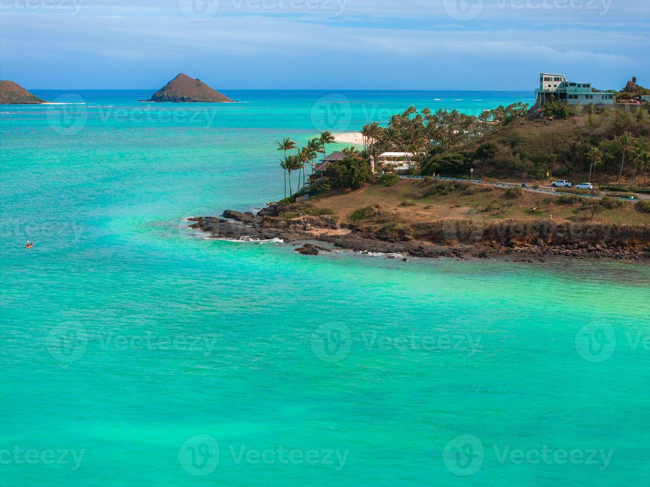 Turquoise ocean waters surround a rocky shoreline on Oahu, Hawaii, with a small offshore islet, palm trees, and a house on a lush hillside. photo