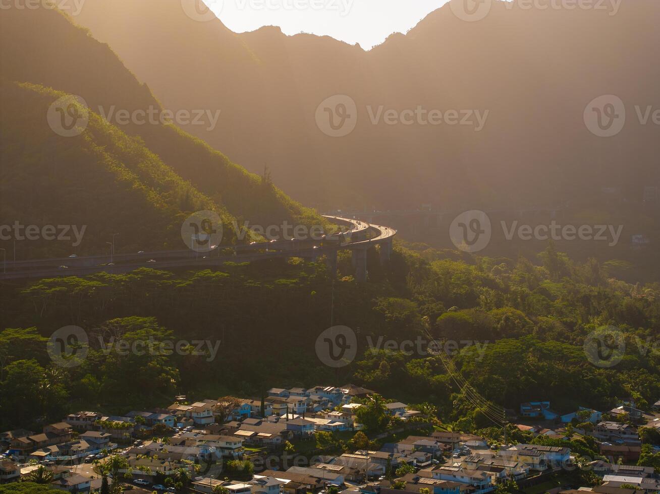 Scenic aerial view of a green valley on Oahu island, Hawaii, with a highway, residential area, and sunlit mountains under golden sunset light. photo