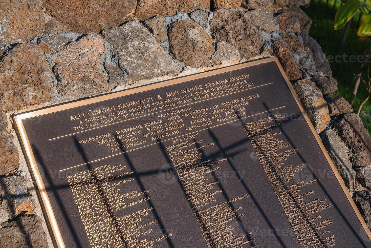 Plaque mounted on a stone structure honoring Ali i Aimoku Kaumuali i and Moi Wahine Kekaiha akulou, surrounded by greenery in an outdoor setting. photo