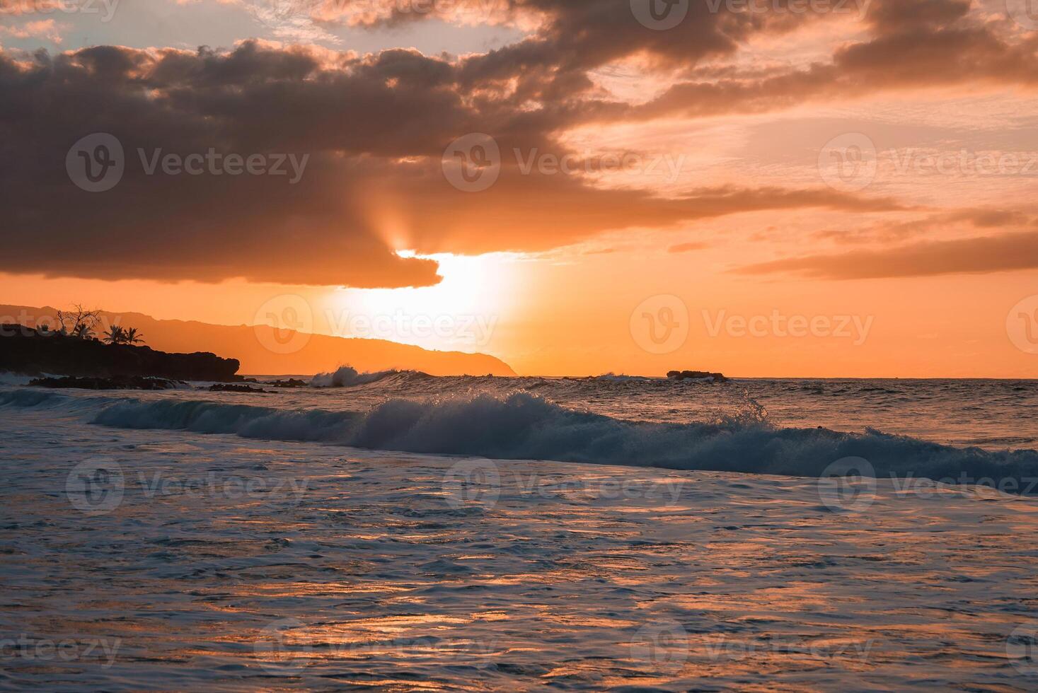 The image shows a sunset over the ocean with warm orange hues, dramatic clouds, rolling waves, a rocky coastline, and silhouetted vegetation. photo