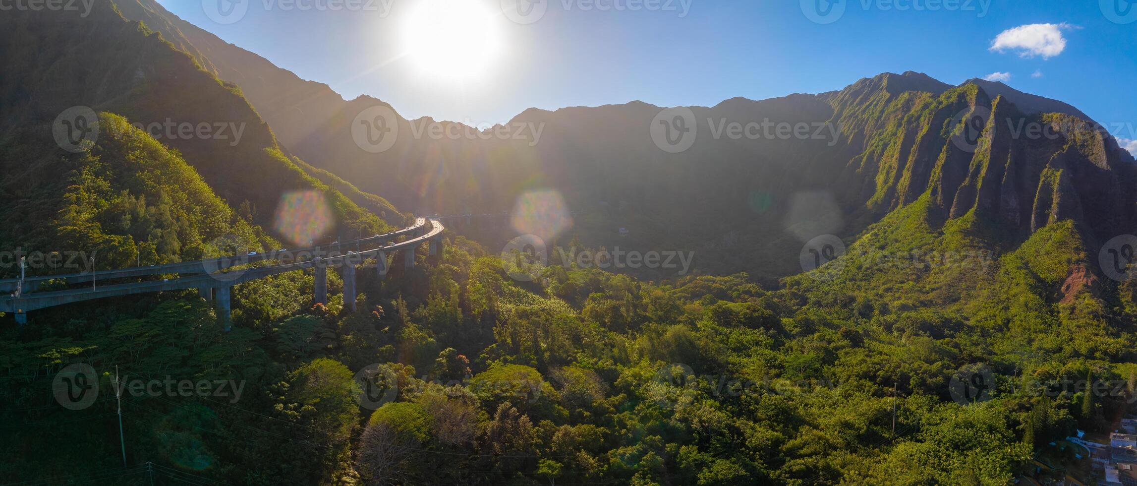 Aerial view of a green valley on Oahu island, Hawaii, with jagged mountain ridges, a curving elevated highway, and sunlight casting a warm glow. photo
