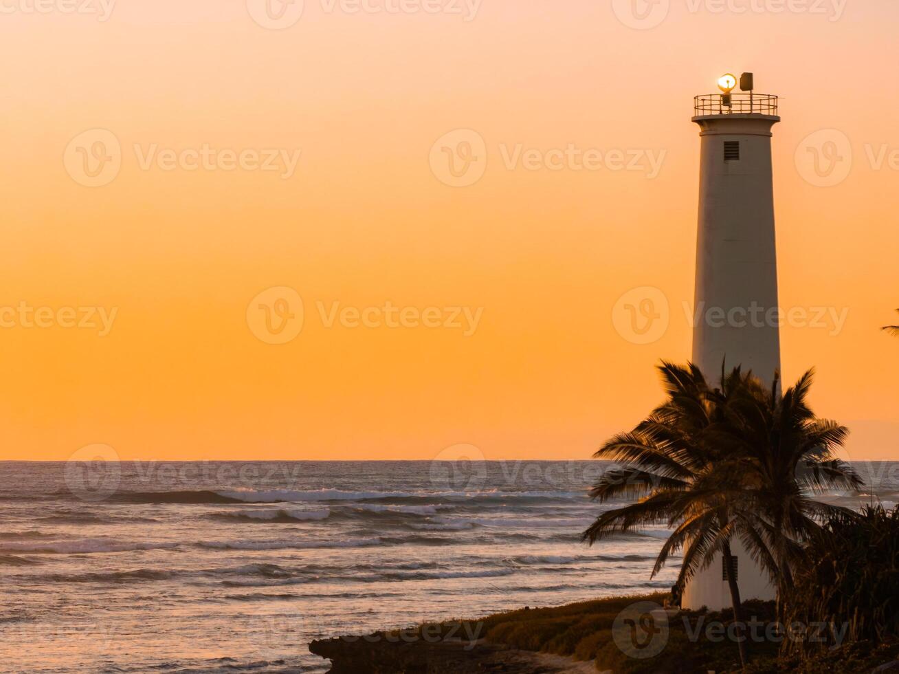 A tall white lighthouse on a coastal cliff, framed by palm trees, overlooks the ocean at sunset. The sky glows orange, contrasting with the blue water. photo