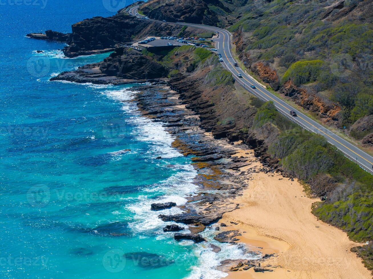 A winding road runs along a rugged cliffside in Oahu, Hawaii, with turquoise ocean waters, a rocky shoreline, and a golden sandy beach below. photo