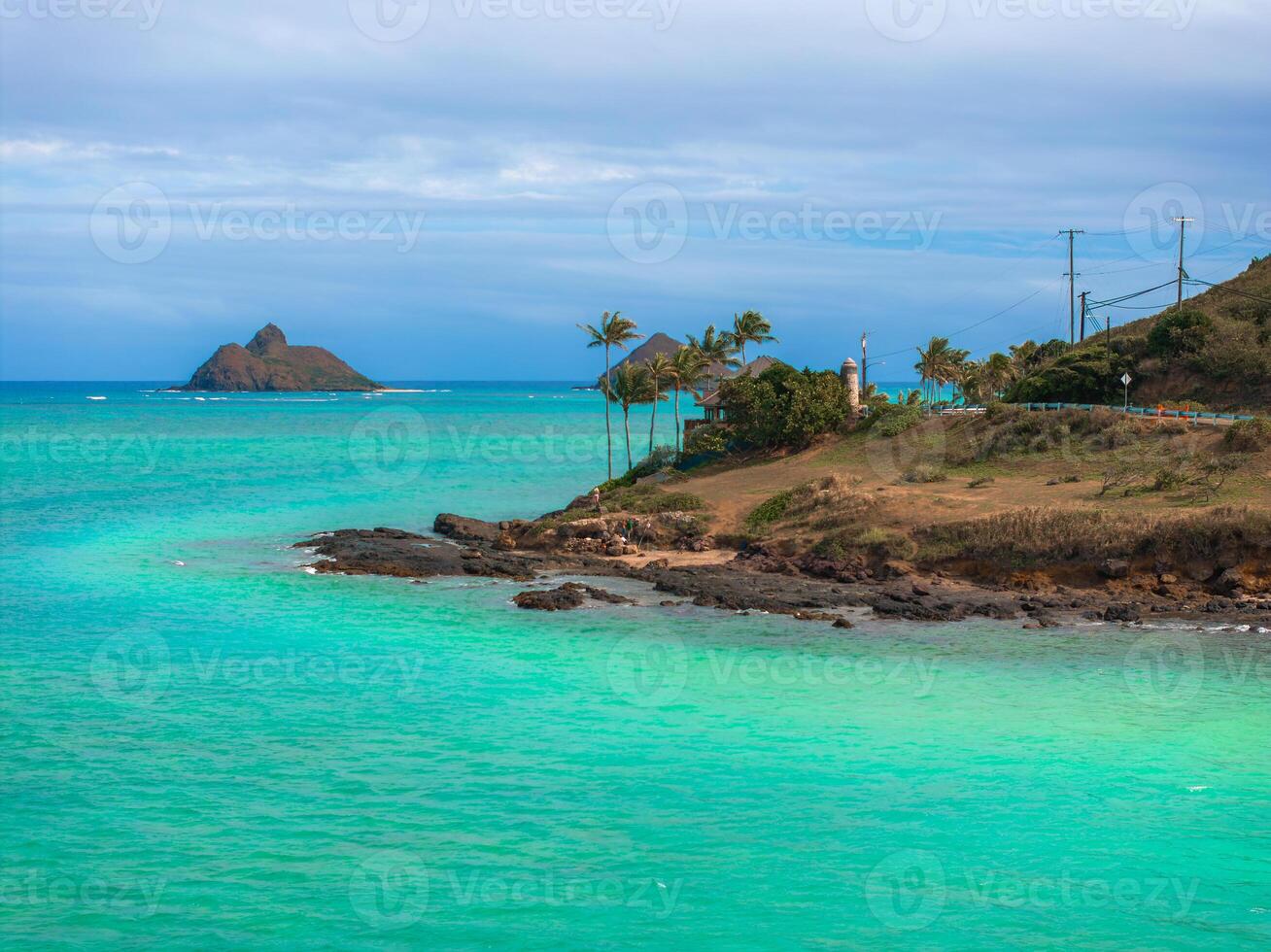 Turquoise waters, rocky shoreline, Mokoli'i Island, palm trees, a lighthouse, power lines, and a road under a partly cloudy sky in Oahu, Hawaii. photo