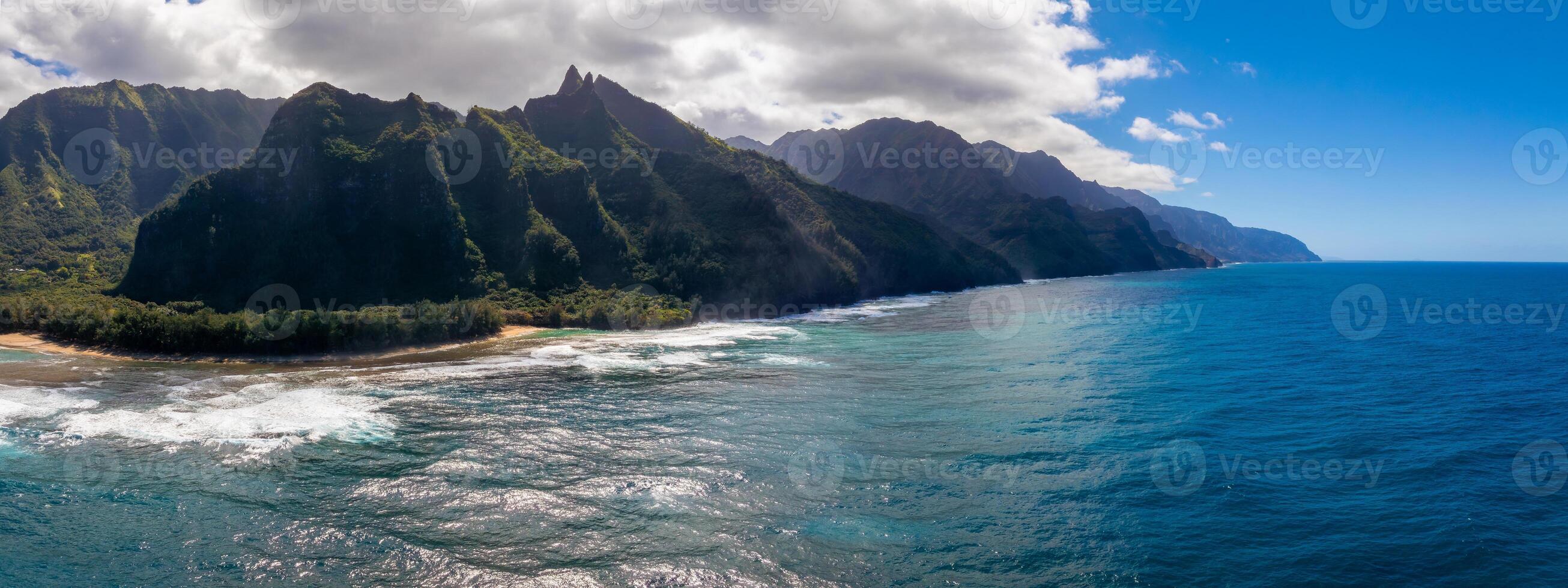 Aerial view of the Napali Coast on Kauai Island, Hawaii, with jagged green cliffs, turquoise waters, white sandy beaches, and scattered clouds. photo
