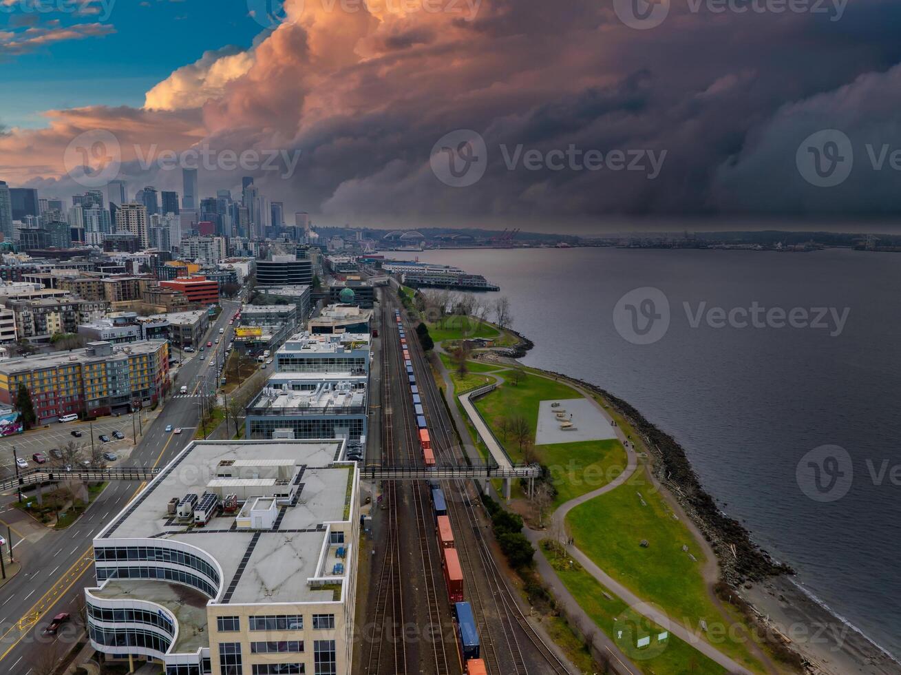 Aerial view of Seattle's downtown skyline, train tracks by a waterfront park, and the bay under a dramatic sky with vibrant and stormy clouds. photo