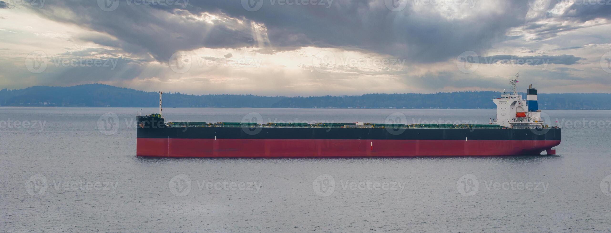 A large cargo ship with a red hull floats on calm waters near a tree lined shoreline. Sunlight breaks through scattered clouds, creating a serene scene. photo
