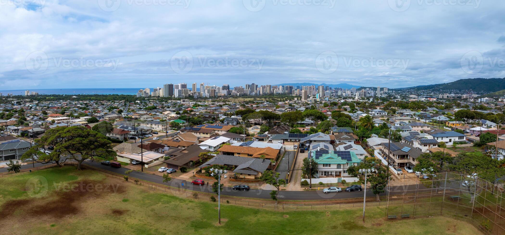 Aerial view of a residential area in Honolulu, Hawaii, with homes, a park, and greenery. The skyline and Pacific Ocean are visible, framed by mountains. photo