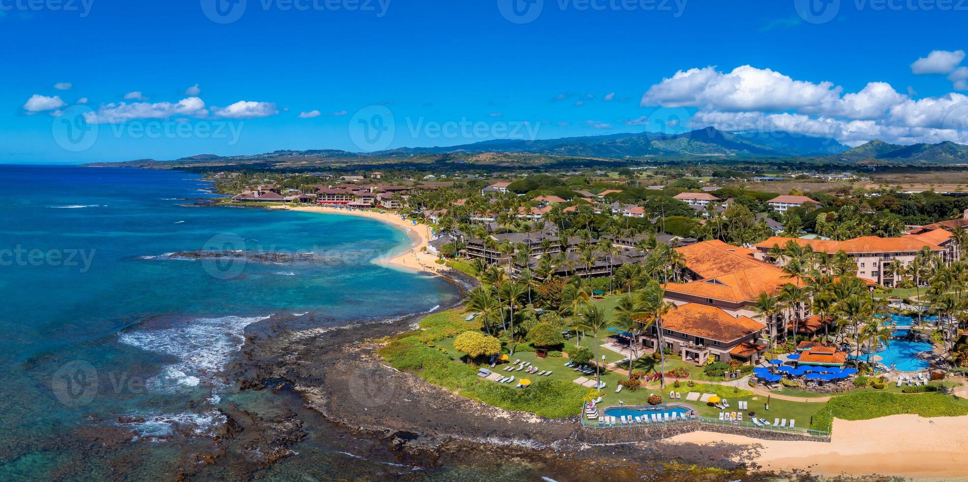 Aerial image of a Kauai island resort with orange tiled roofs, a pool, palm trees, a sandy beach, turquoise ocean, and distant green mountains. photo