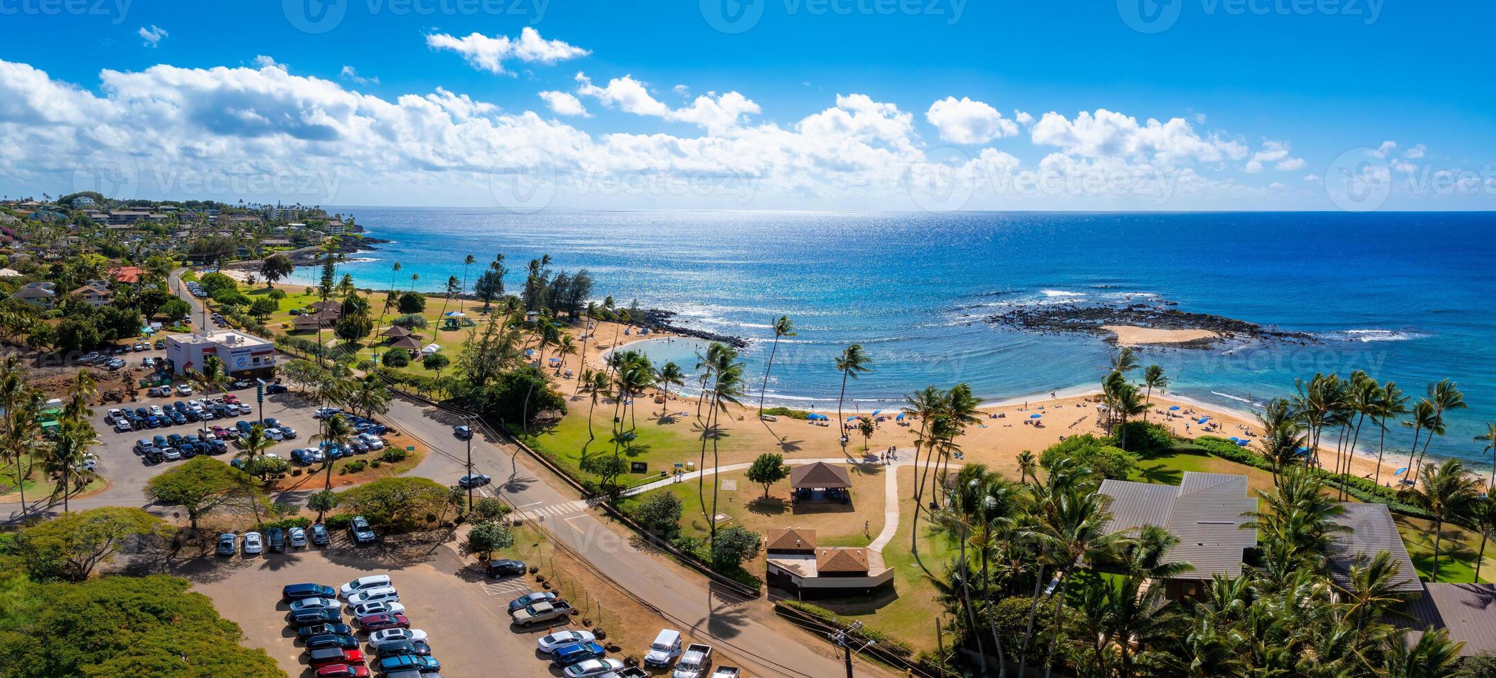 Aerial view of a golden sandy beach on Kauai island, Hawaii, bordered by turquoise waters, rocky outcrops, palm trees, and a nearby park with pathways. photo