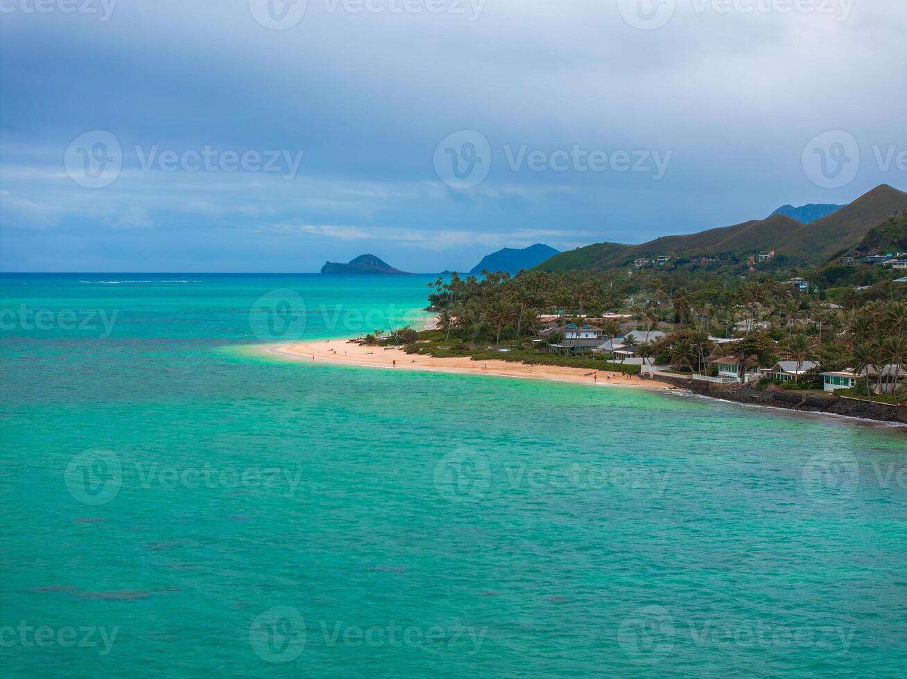 Turquoise ocean meets sandy beach lined with palm trees and homes, with lush hills and a small offshore island under a cloudy sky in Oahu, Hawaii. photo