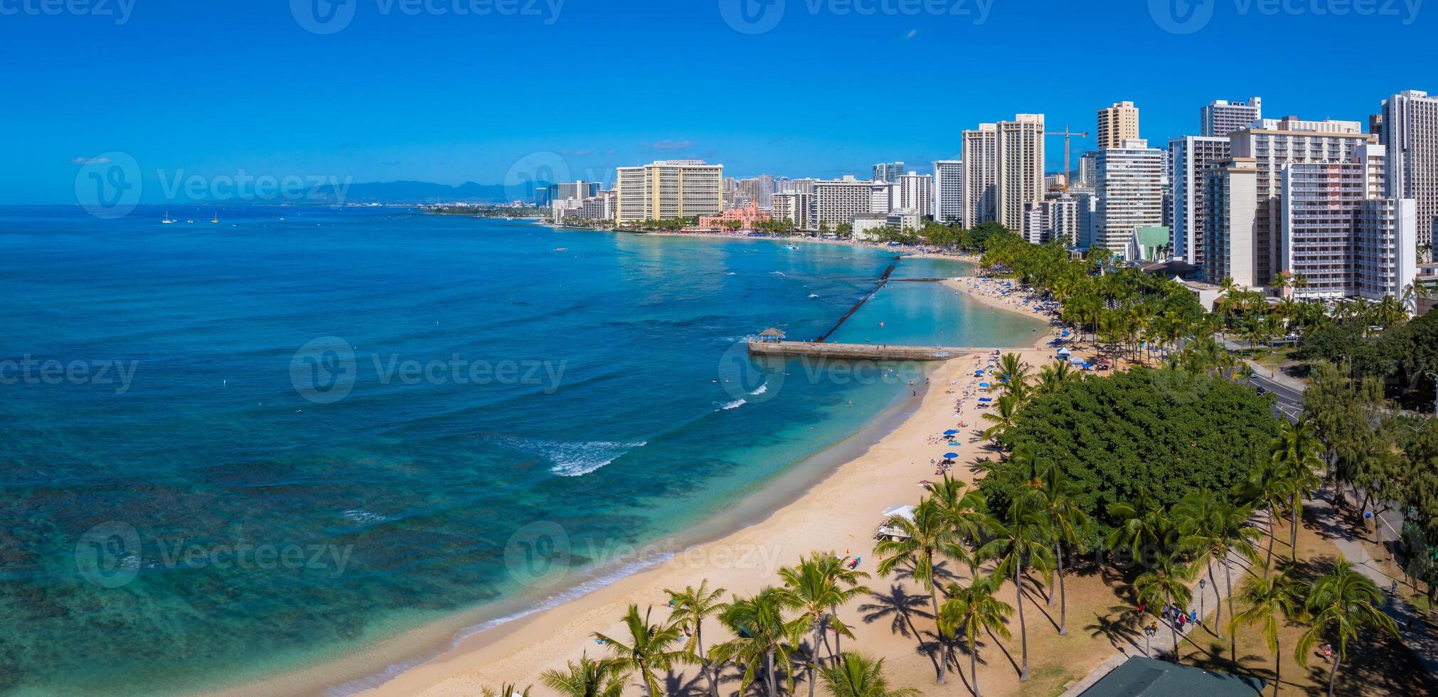 Turquoise waters meet sandy shores at Waikiki Beach, with palm trees, a jetty, and high rise buildings under a clear blue sky in Honolulu, Oahu. photo