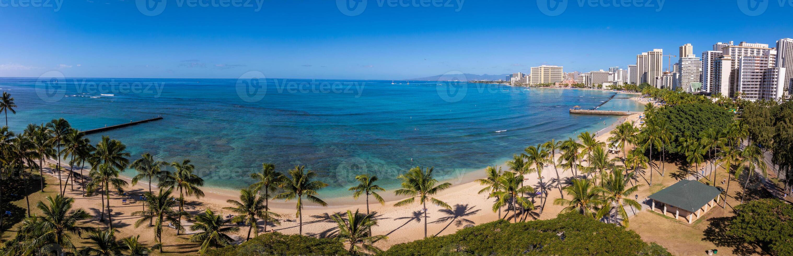 Aerial view of Waikiki Beach in Honolulu, Oahu, Hawaii, featuring turquoise waters, sandy shores, palm trees, piers, and high rise buildings under a clear sky. photo