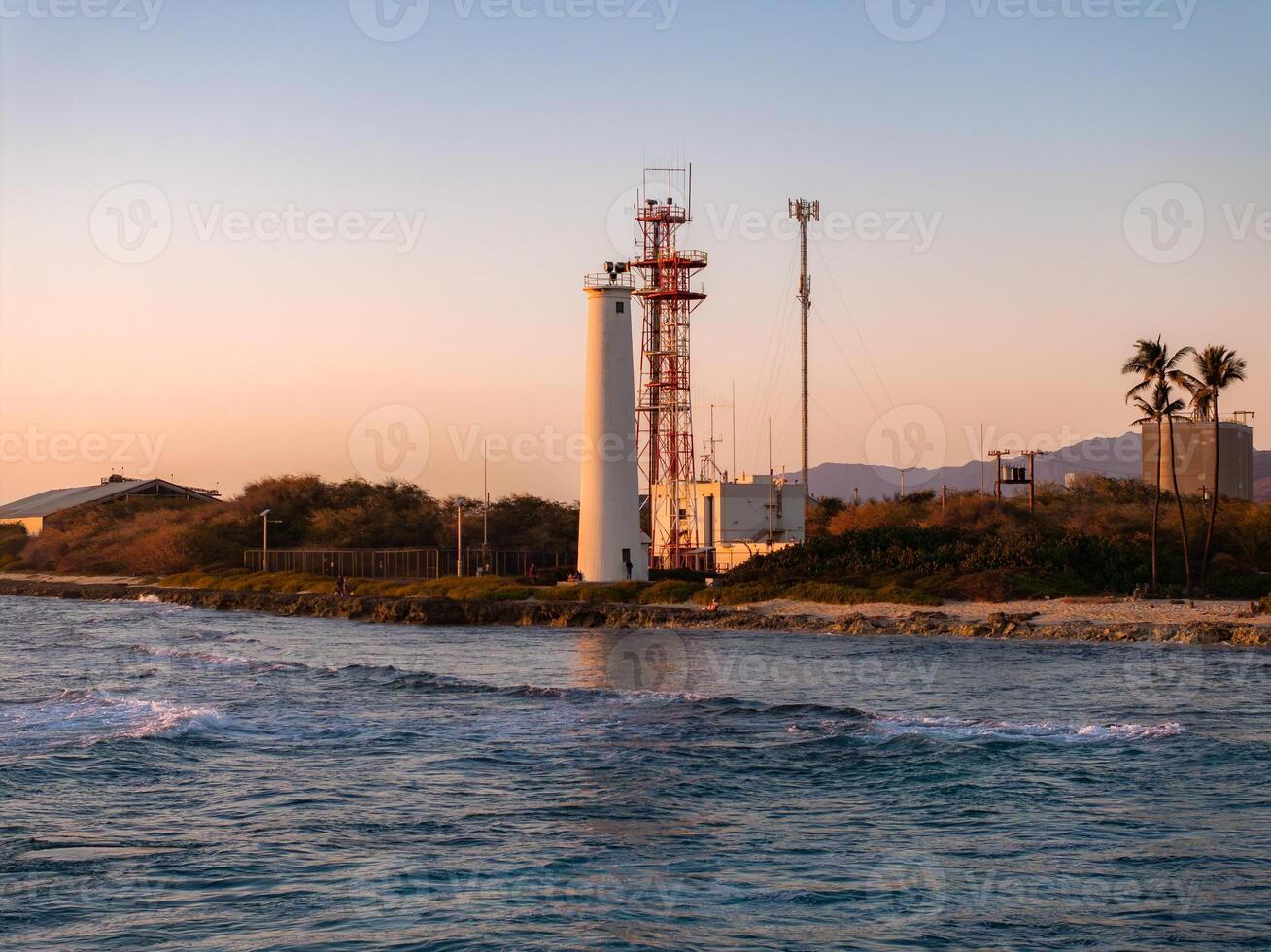 A white lighthouse with red accents near a rocky shoreline at sunset, surrounded by sparse vegetation, palm trees, and distant mountains on Oahu. photo