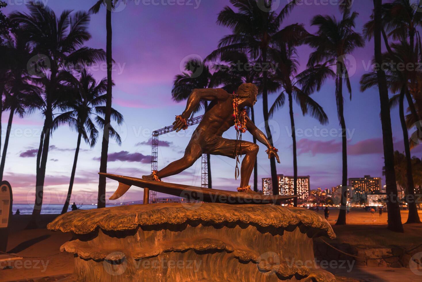 The bronze Duke Kahanamoku statue adorned with flower leis, set against palm trees, a vibrant sunset sky, and Waikiki's illuminated cityscape. photo