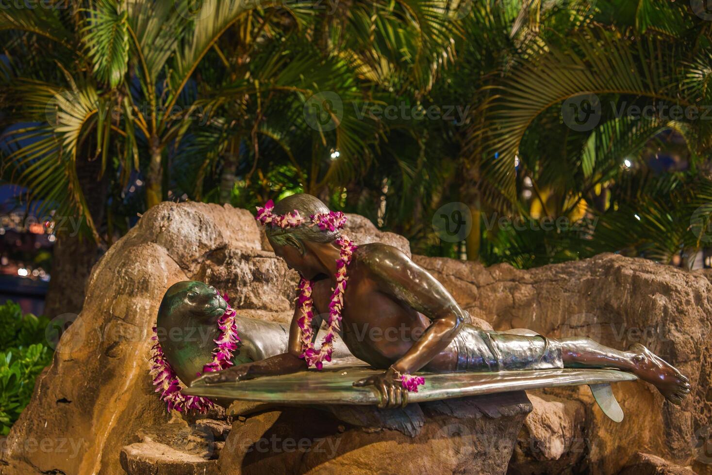 A bronze statue of a surfer on a surfboard, adorned with Hawaiian flower leis, set against tropical plants and rocks under soft nighttime lighting. photo
