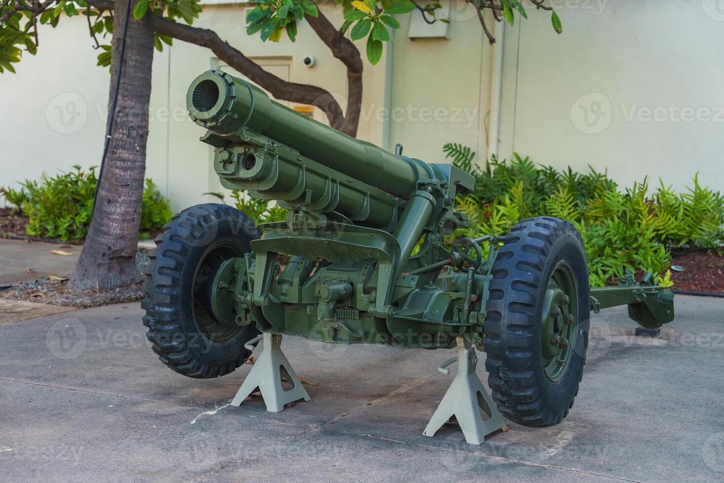 A green military artillery cannon rests on white stands outdoors, surrounded by tropical vegetation and a light colored building wall in Hawaii. photo