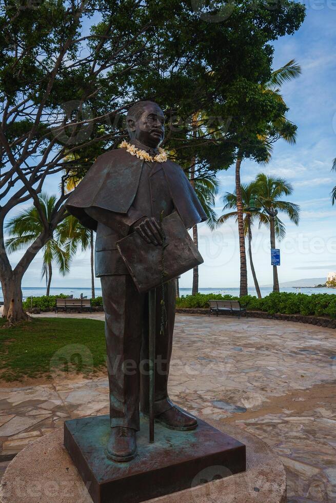 A bronze statue of a man with a lei stands on a stone pedestal in a park with palm trees, benches, and an ocean view, surrounded by tropical greenery. photo