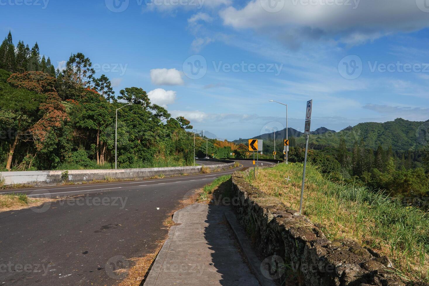 A scenic road bordered by greenery and a stone wall curves left, with hills and mountains in the background under a clear sky with scattered clouds. photo