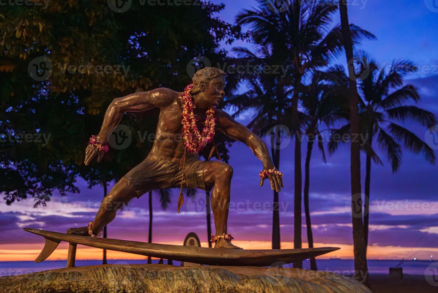 A bronze surfer statue adorned with flower leis stands on a surfboard, framed by palm trees, a vibrant sunset, and a serene Hawaiian beach. photo