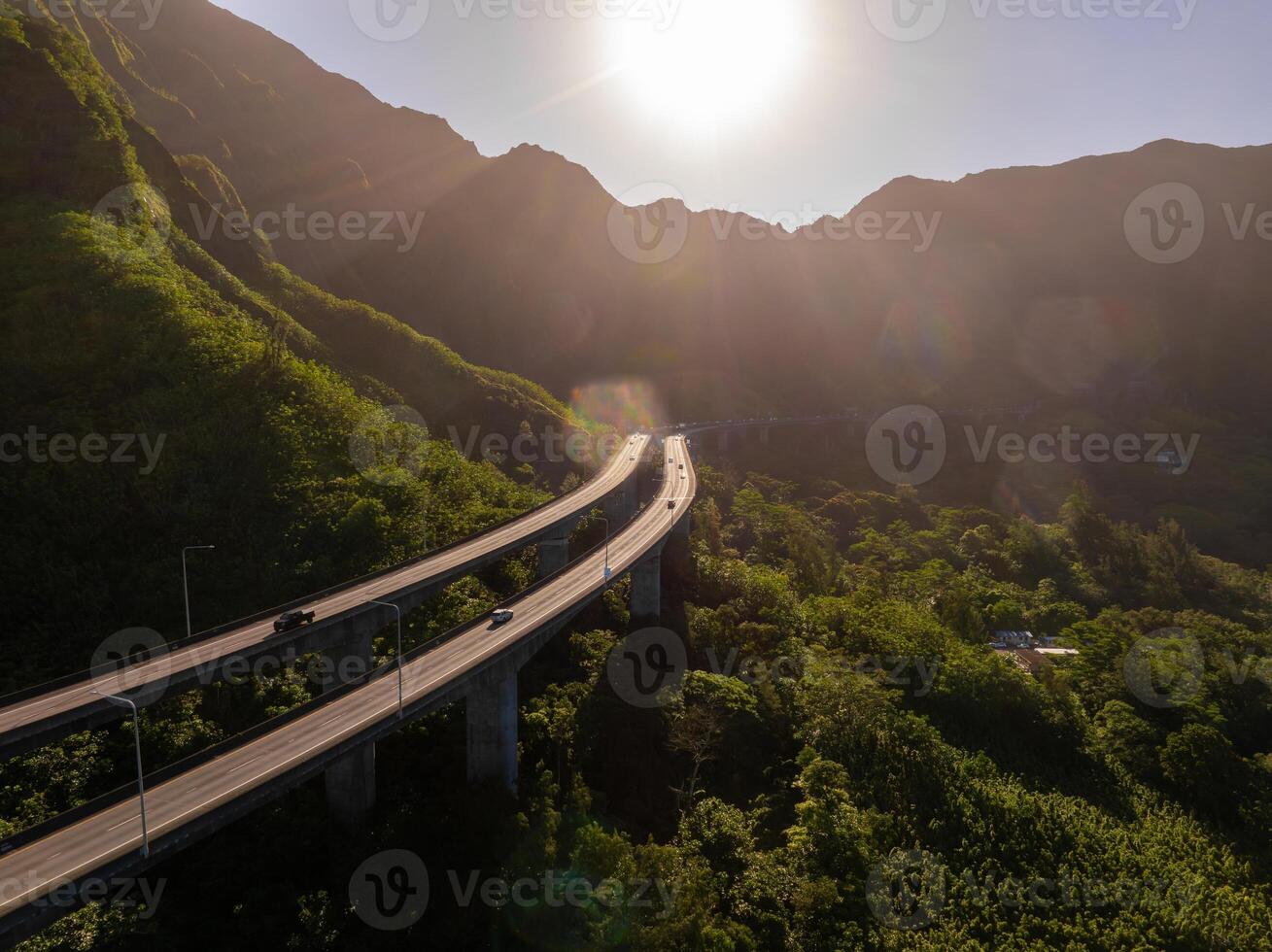 An elevated highway curves through green mountains on Oahu, Hawaii, with sunlight illuminating tropical vegetation and dramatic peaks in the background. photo