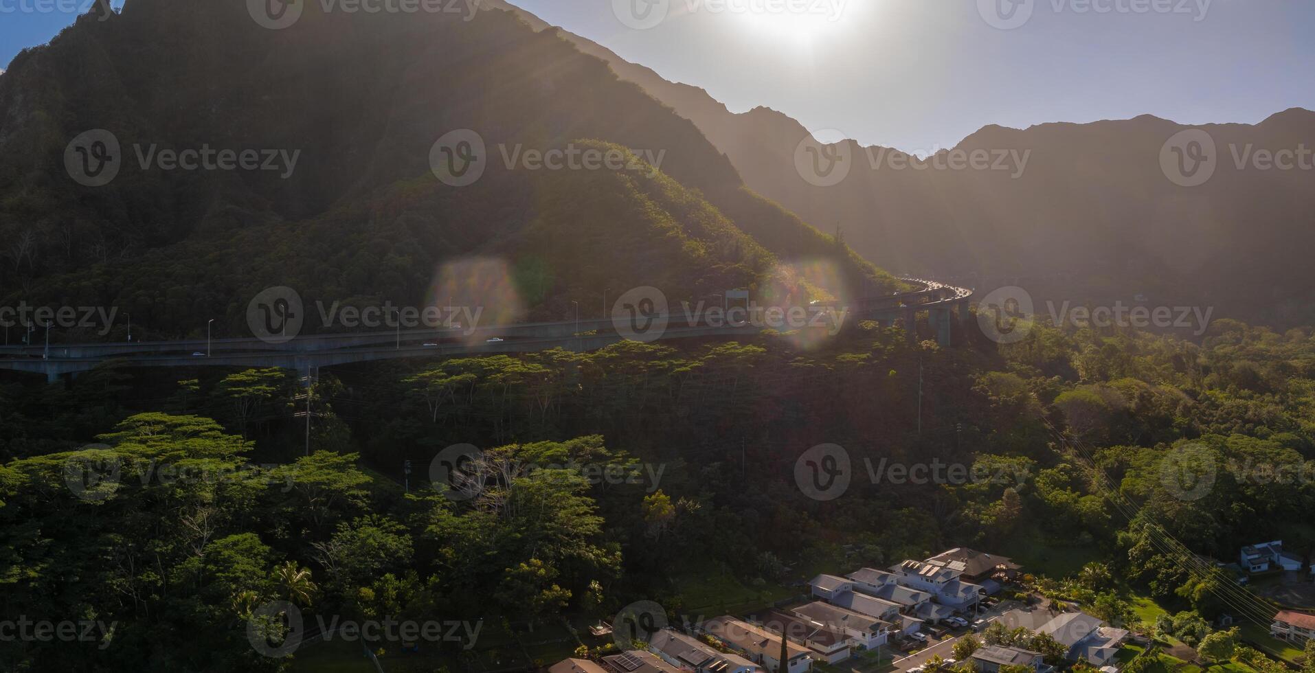 Aerial view of green mountains on Oahu island, Hawaii, with a highway curving above tropical vegetation and a small residential area below. photo
