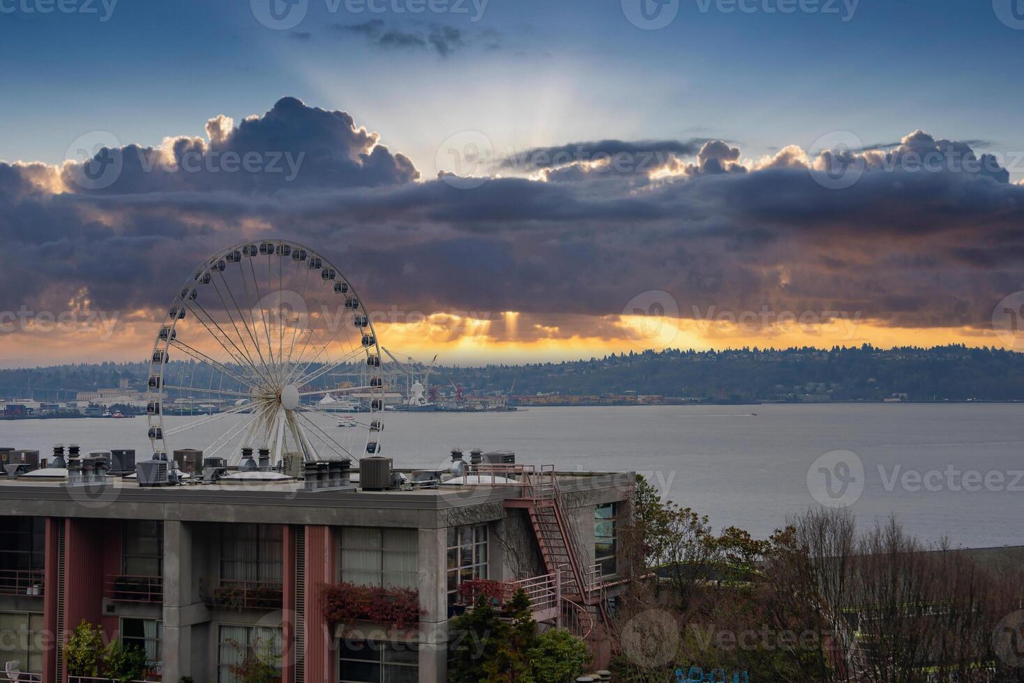 The Seattle Great Wheel stands on the waterfront as golden sunset rays pierce thick clouds. Foreground shows a building with greenery, and Elliott Bay lies beyond. photo