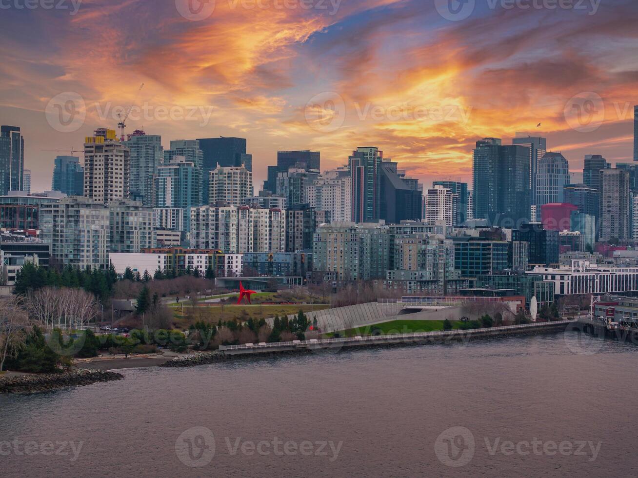 Aerial view of Seattle's downtown skyline at sunset, featuring high rise buildings, a red sculpture in a waterfront park, and calm bay waters. photo