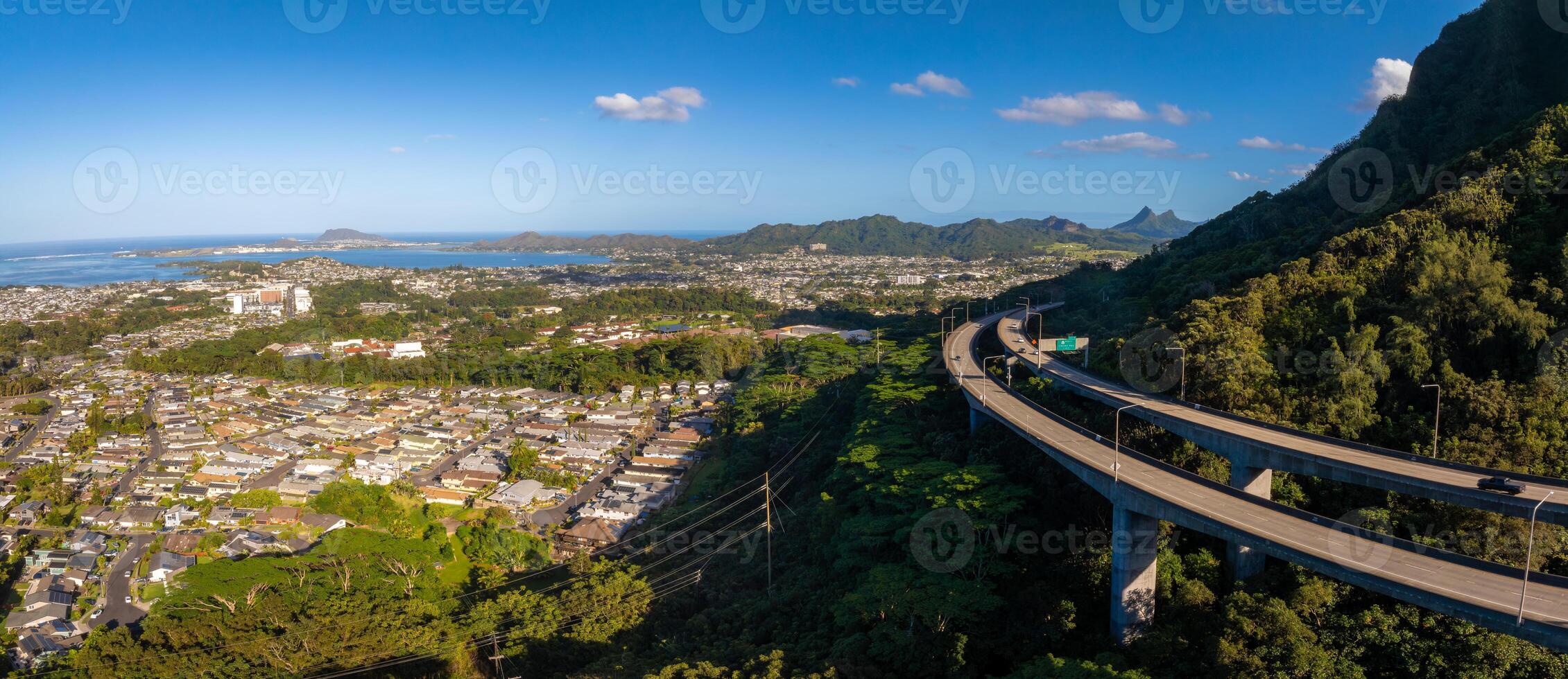 Aerial view of Oahu, Hawaii, showing a highway through green hills, a residential area, rugged mountains, and a distant coastline under bright daylight. photo
