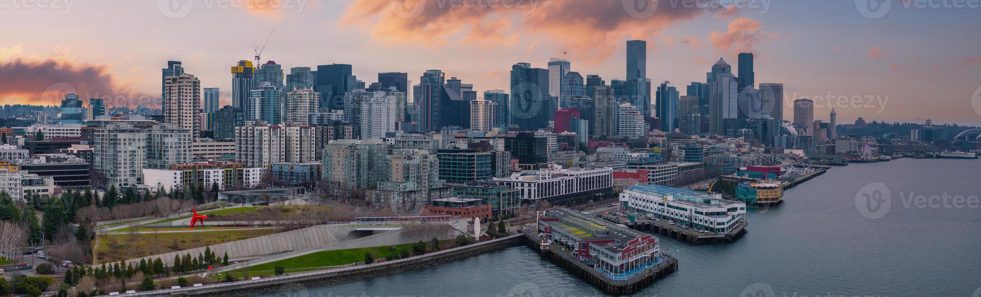 Downtown Seattle skyline with the Space Needle, waterfront piers, a cargo ship, the Seattle Great Wheel, and a park with a red sculpture at sunset. photo