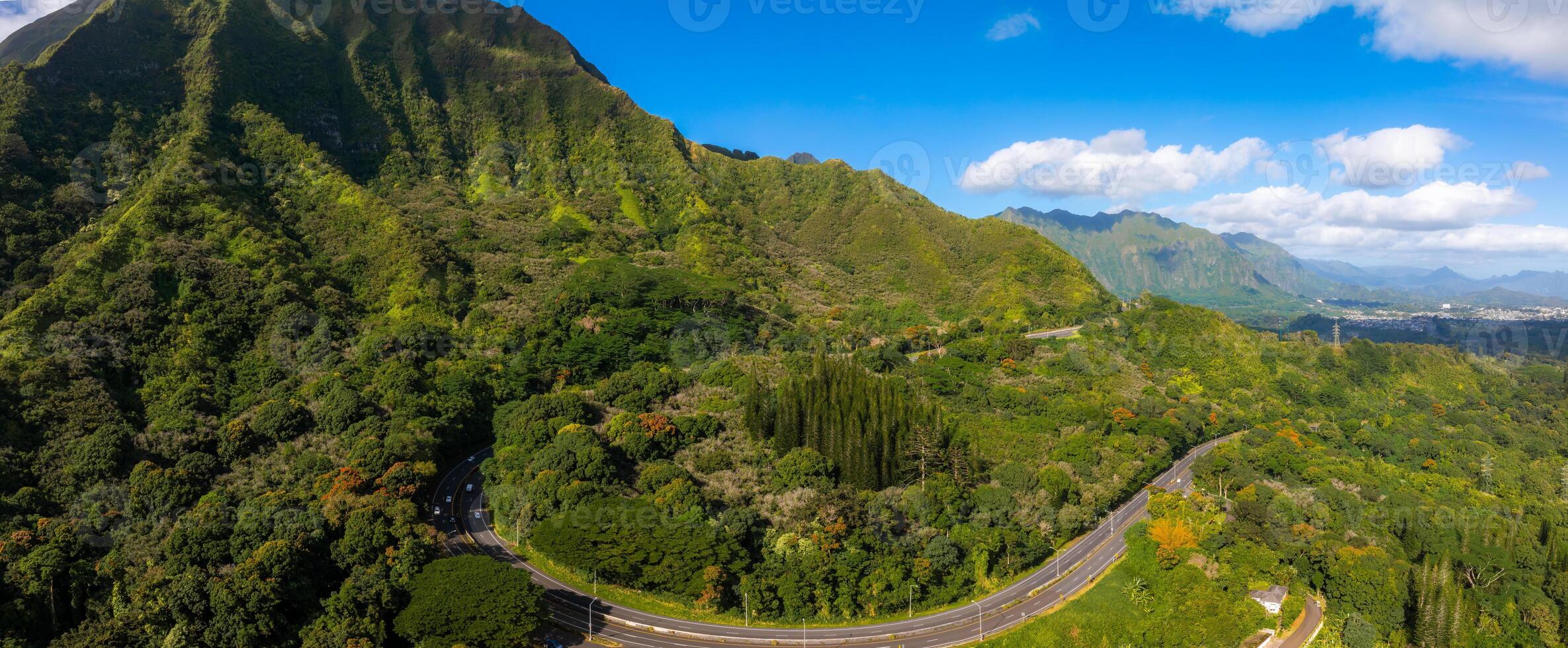 Aerial view of green mountains and valleys on Oahu Island, Hawaii, with a winding highway cutting through dense tropical vegetation under a clear sky. photo