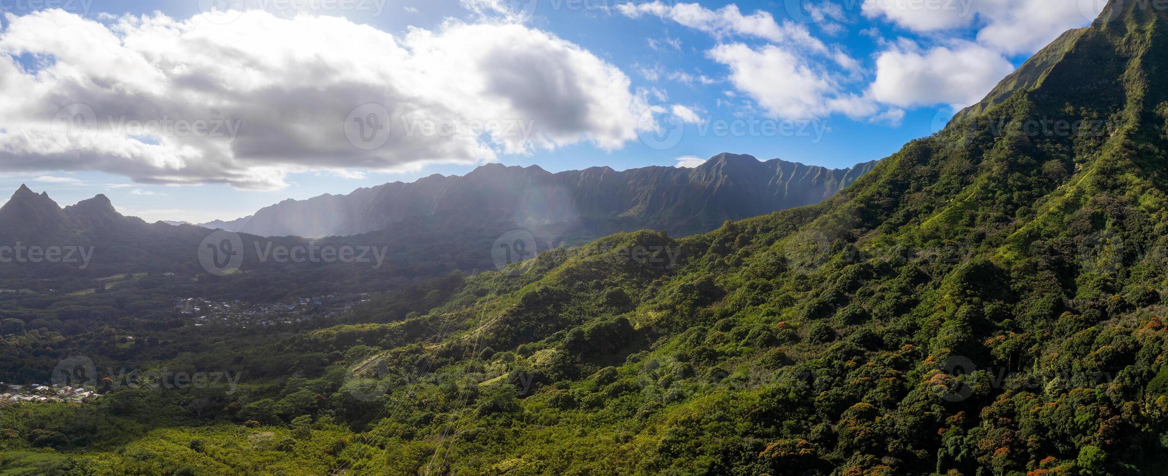 Lush green mountains and valleys of the Ko olau Range on Oahu Island, Hawaii, with sunlight filtering through clouds and a settlement in the valley. photo