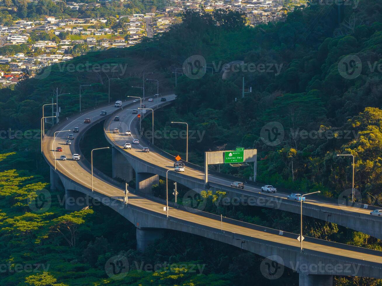 An elevated highway curves through dense vegetation on Oahu island, Hawaii, with a residential area in the background and a green road sign visible. photo