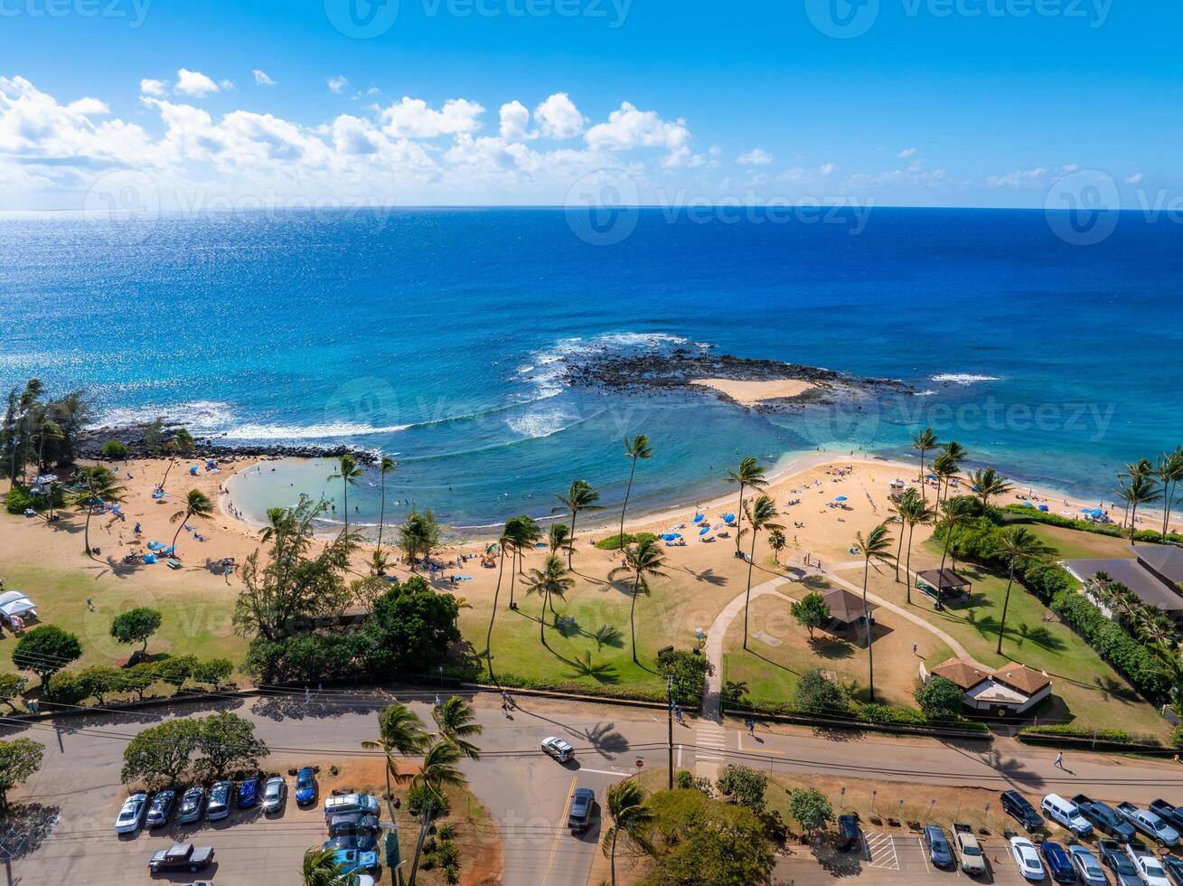 Aerial image of a Kauai beach in Hawaii featuring golden sand, turquoise waters, palm trees, umbrellas, visitors, a parking lot, and nearby buildings. photo