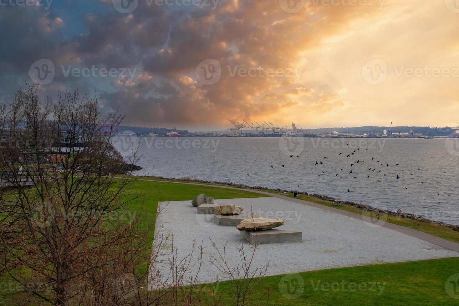 A waterfront park in Seattle, USA, during sunset features stone sculptures on gravel, bare trees, industrial cranes, and birds in flight over the bay. photo