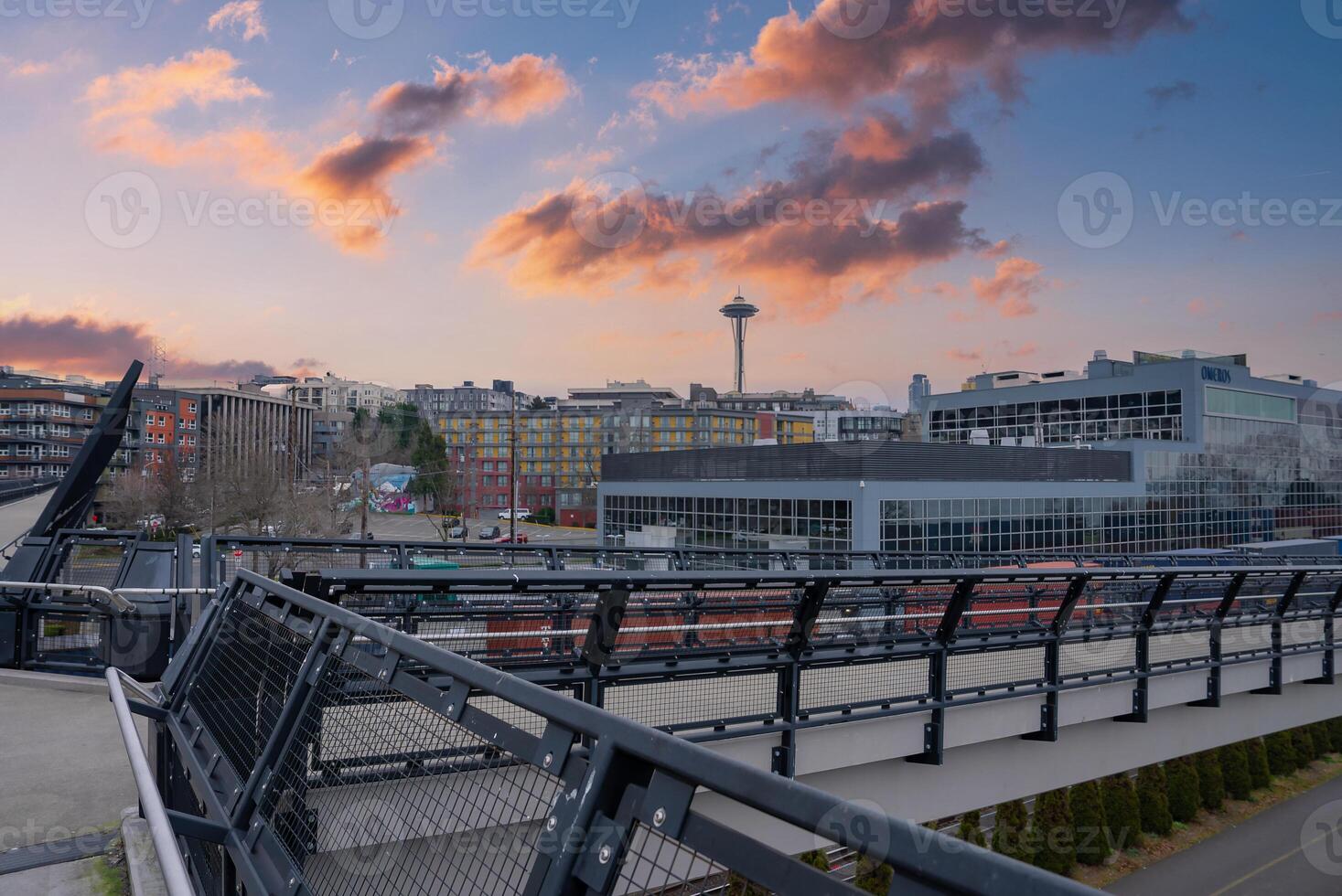 View of Seattle with the Space Needle, a modern pedestrian bridge, colorful buildings, and greenery under a vibrant sunset sky with scattered clouds. photo