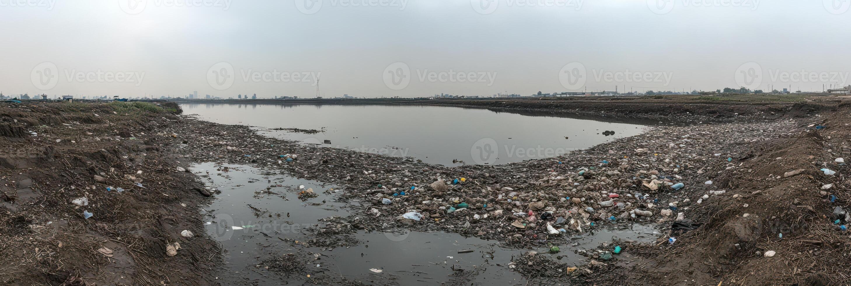 Polluted riverbank covered with trash and debris in urban environment under cloudy sky photo