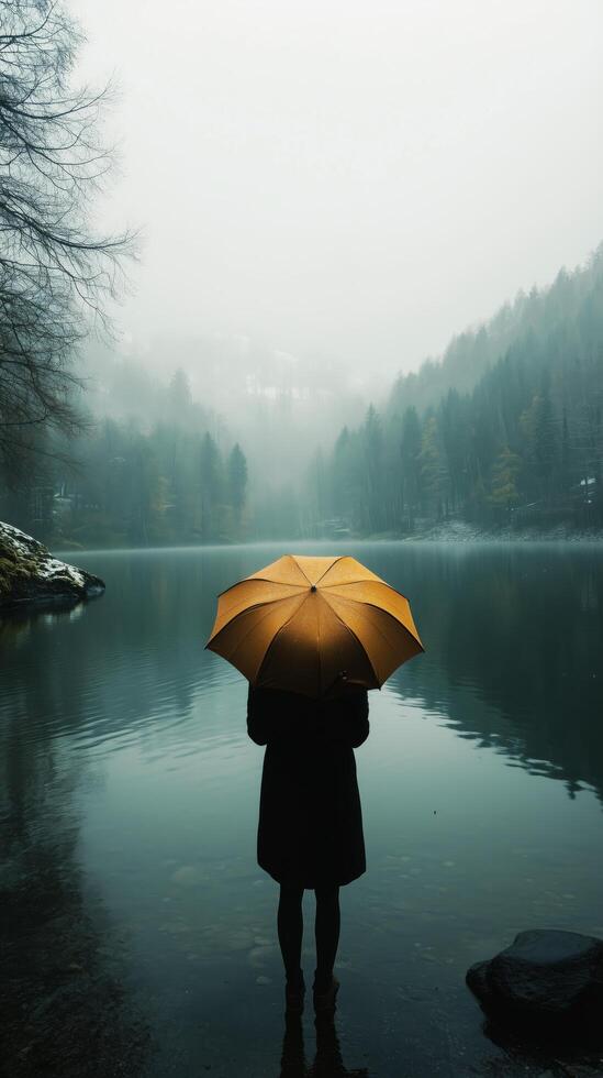 Person with yellow umbrella stands by tranquil lake in misty forest during early morning. photo