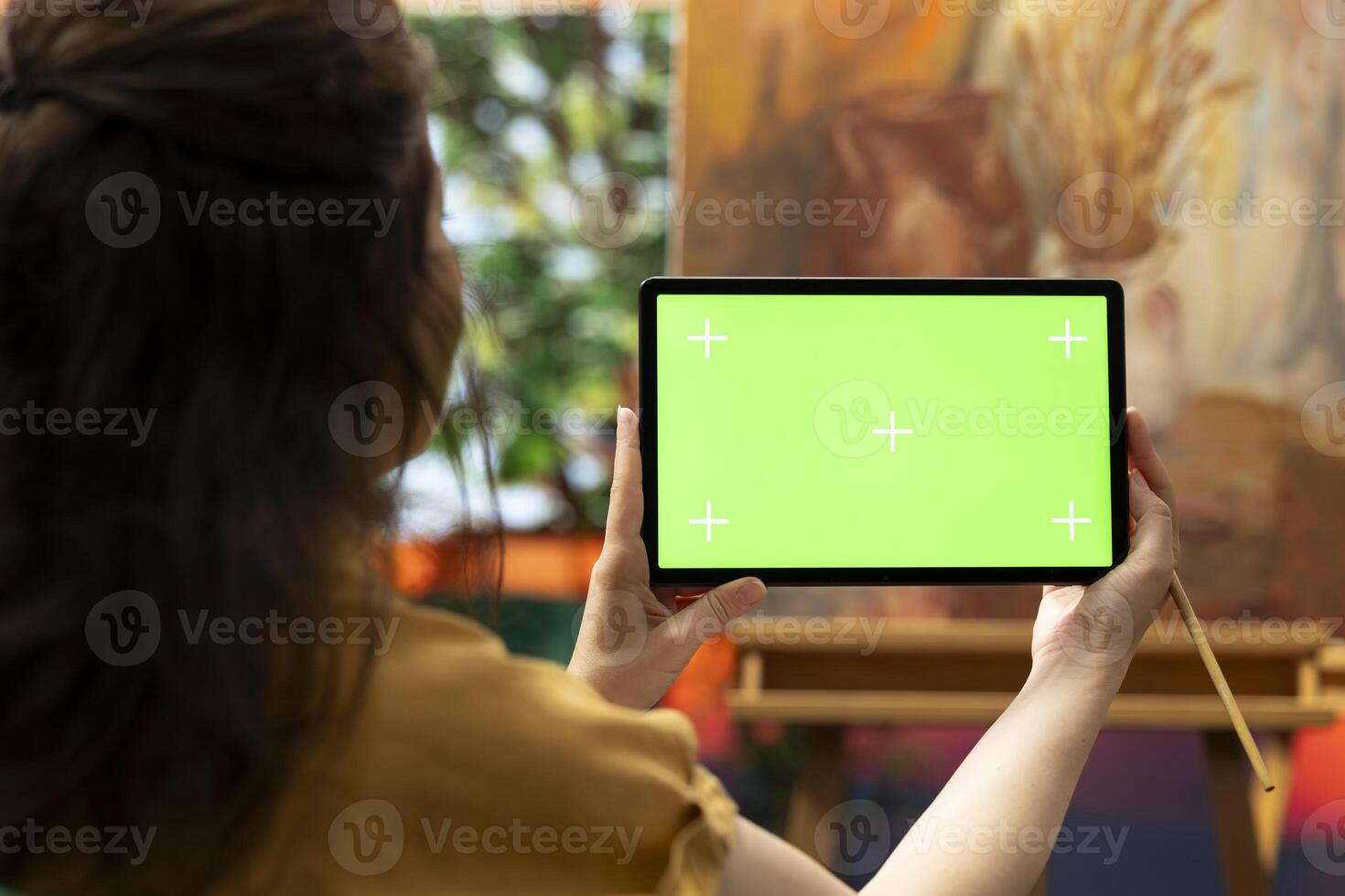 Painter holding a device with isolated mockup screen in her art studio, looking at her finished masterpiece with oil paints on canvas. Beautiful rustic artwork for a fine art gallery, creativity. photo