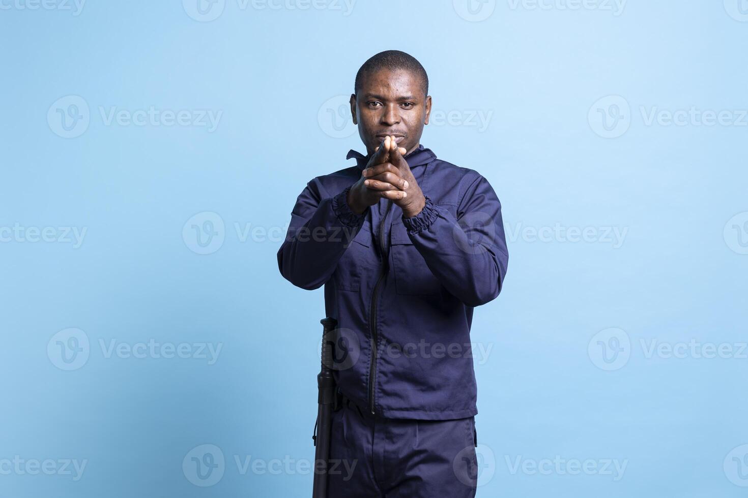 African american security agent mimicking target shooting with an imaginary gun, holding his hands in the shape of a pistol. Confident bodyguard acting serious against blue background. photo