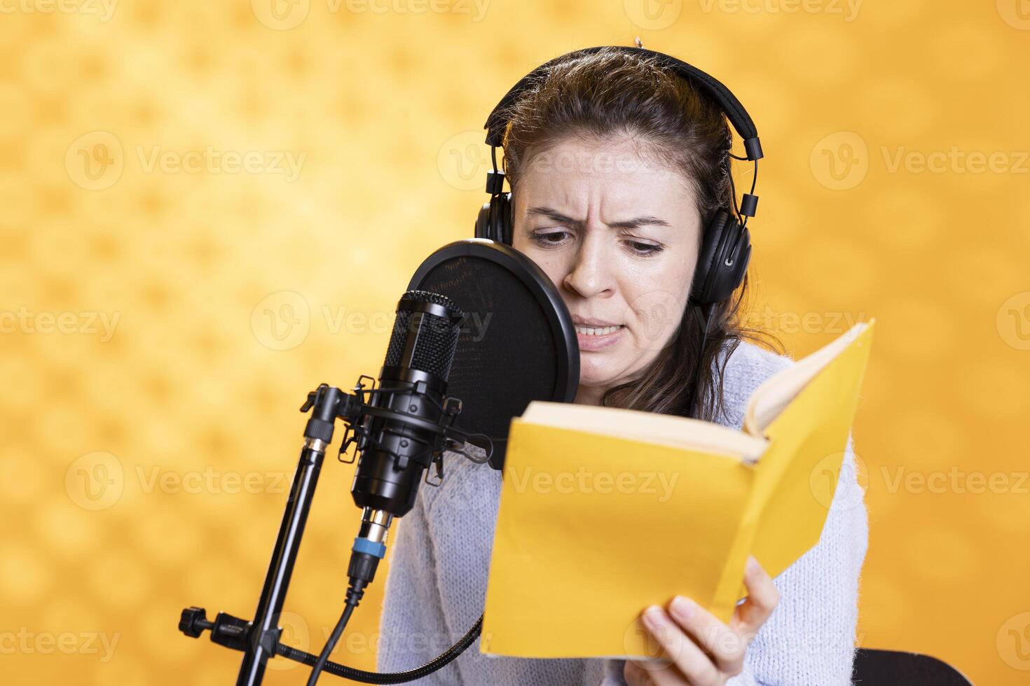 Narrator frowning while reading book, recording audiobook using microphone, playing angry character, studio background. Woman producing digital recording of novel, interpreting furious antagonist photo
