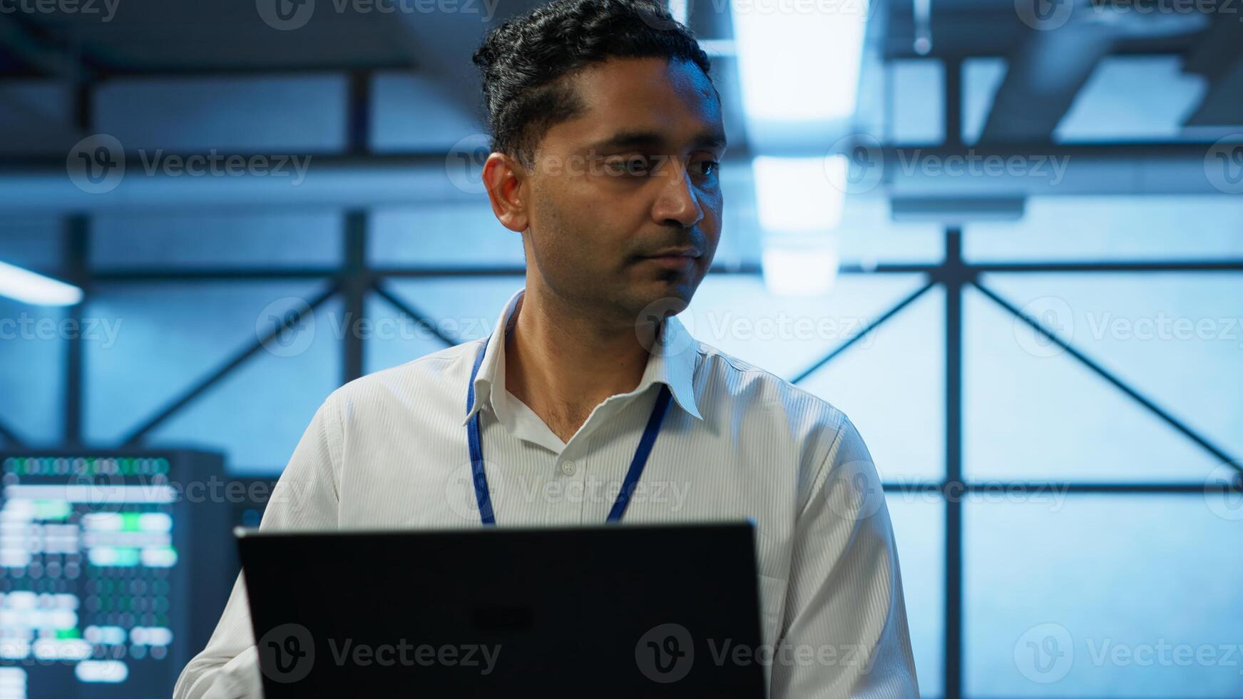 System administrator in data center using notebook to design, implement and manage gear, ensuring optimal performance and reliability. Indian man in server room coding on laptop, camera A photo