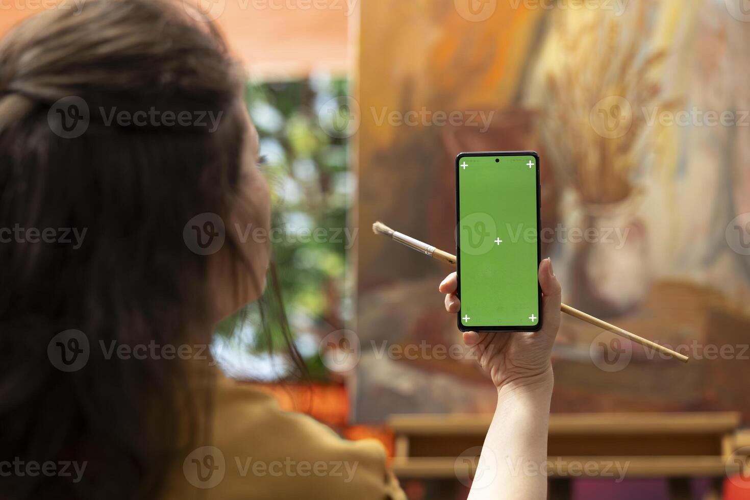 Painter holding a device with isolated mockup screen in her art studio, looking at her finished masterpiece with oil paints on canvas. Beautiful rustic artwork for a fine art gallery, creativity. photo