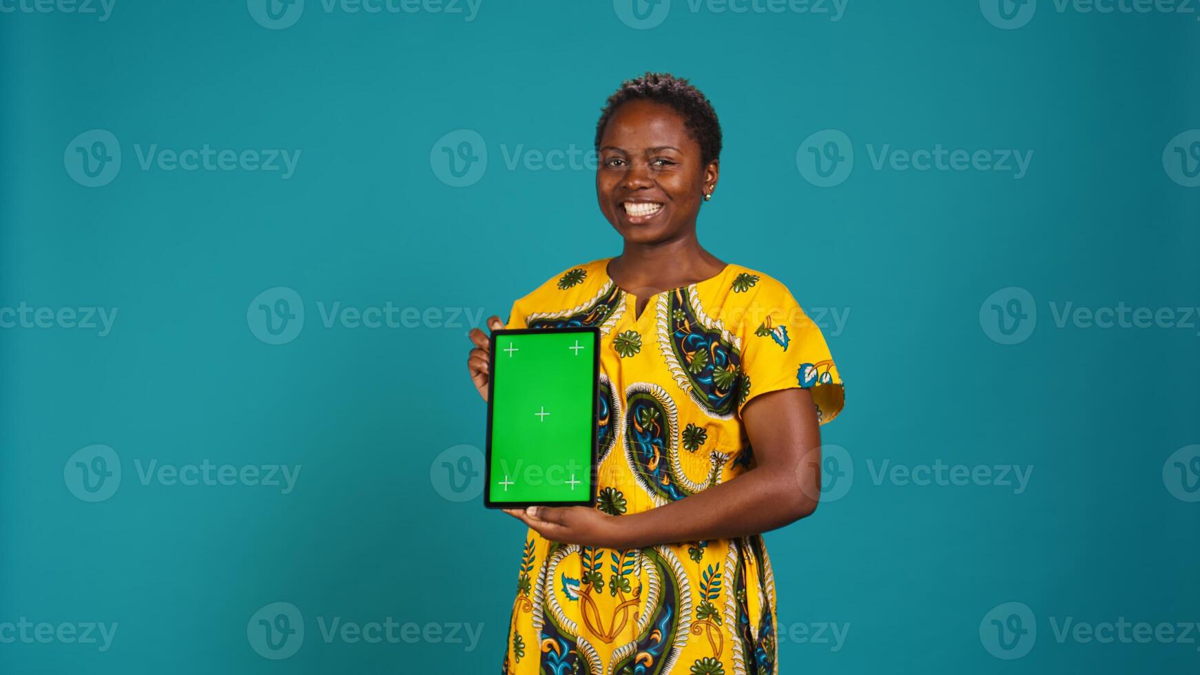 Gorgeous model presenting a tablet with green screen mockup, holding the gadget against blue background. Young charming woman in tribal clothing showing isolated display in studio. Camera B. photo