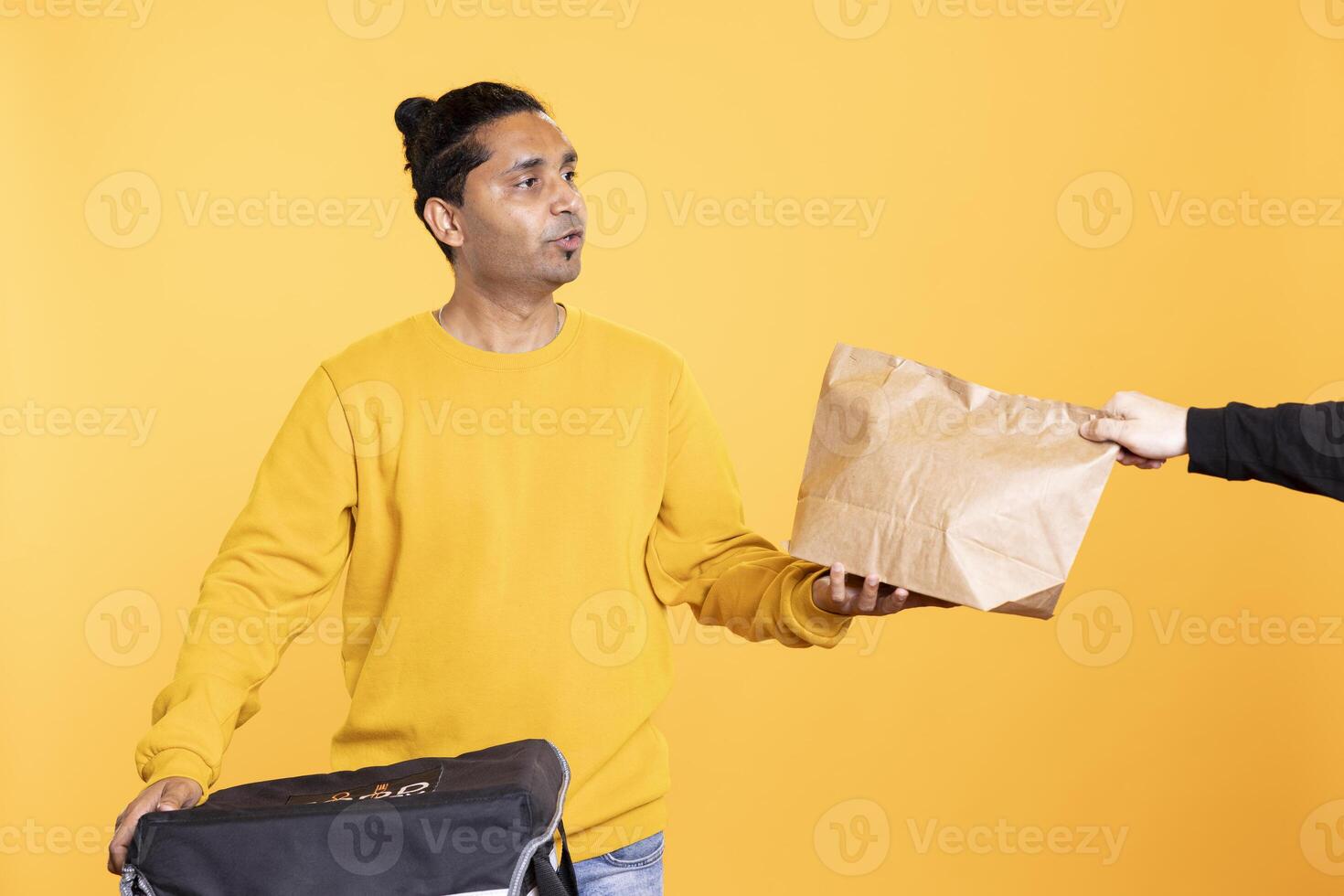Delivery employee getting takeaway order out of thermal backpack, giving it to client. Courier offering lunch paper bag to hungry customer, isolated over studio background photo