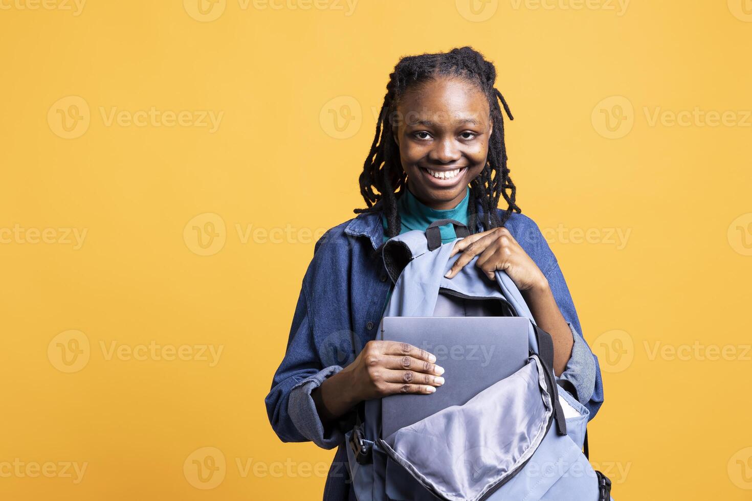 Portrait of happy young girl putting laptop in backpack, preparing to go to school exam, isolated over studio background. Satisfied teenager packing university notebook in rucksack photo